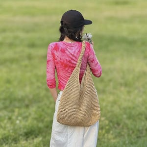 Puede incluir: Un bolso de hombro beige tejido con una correa larga. El bolso se lleva sobre el hombro. La persona de la imagen lleva una gorra de b&eacute;isbol negra, una camisa de manga larga rosa te&ntilde;ida con tinte y pantalones blancos. El fondo es un campo verde.