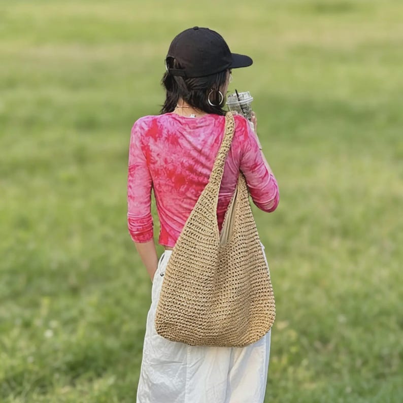 Puede incluir: Un bolso de hombro beige tejido con una correa larga. El bolso se lleva sobre el hombro. La persona de la imagen lleva una gorra de b&eacute;isbol negra, una camisa de manga larga rosa te&ntilde;ida con tinte y pantalones blancos. El fondo es un campo verde.