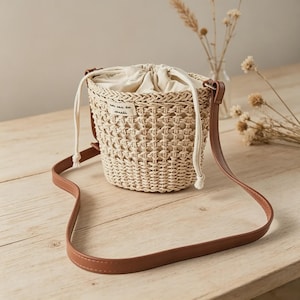 Puede incluir: Un bolso de cubo de paja tejida con cierre de cordón y una correa de hombro de cuero marrón. El bolso tiene un interior color crema y una pequeña etiqueta con texto. El bolso está sobre una superficie de madera.