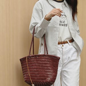 Peut inclure: Un sac fourre-tout bordeaux tiss&eacute; avec des anses tress&eacute;es. Le sac a une forme rectangulaire et une fermeture &agrave; cordon en cuir. Le sac est tenu par une personne portant une chemise ray&eacute;e et un pantalon blanc.