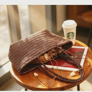 Peut inclure: Un sac fourre-tout tiss&eacute; marron avec des poign&eacute;es tress&eacute;es et des accents en bois est pos&eacute; sur une table en bois. Le sac est ouvert, r&eacute;v&eacute;lant une fermeture &agrave; cordon. Une tasse &agrave; caf&eacute; blanche avec un logo vert et un magazine sont &eacute;galement sur la table.