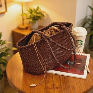 Peut inclure: Un sac fourre-tout marron tress&eacute; avec une poign&eacute;e tress&eacute;e et une fermeture &agrave; cordon. Le sac est fait d'un mat&eacute;riau marron fonc&eacute; avec un motif de tissage en panier. Le sac est sur une table en bois.