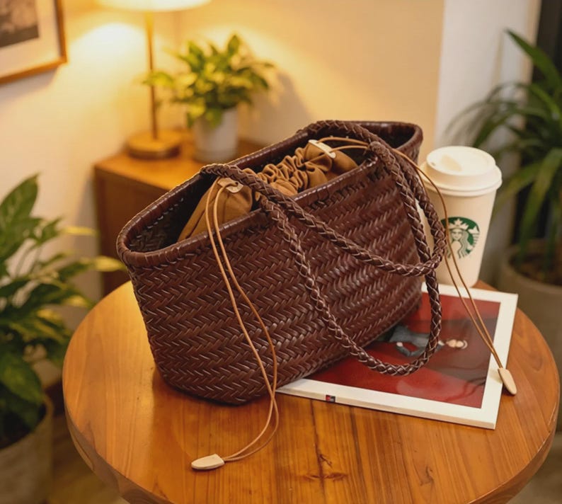 Peut inclure: Un sac fourre-tout marron tress&eacute; avec une poign&eacute;e tress&eacute;e et une fermeture &agrave; cordon. Le sac est fait d'un mat&eacute;riau marron fonc&eacute; avec un motif de tissage en panier. Le sac est sur une table en bois.