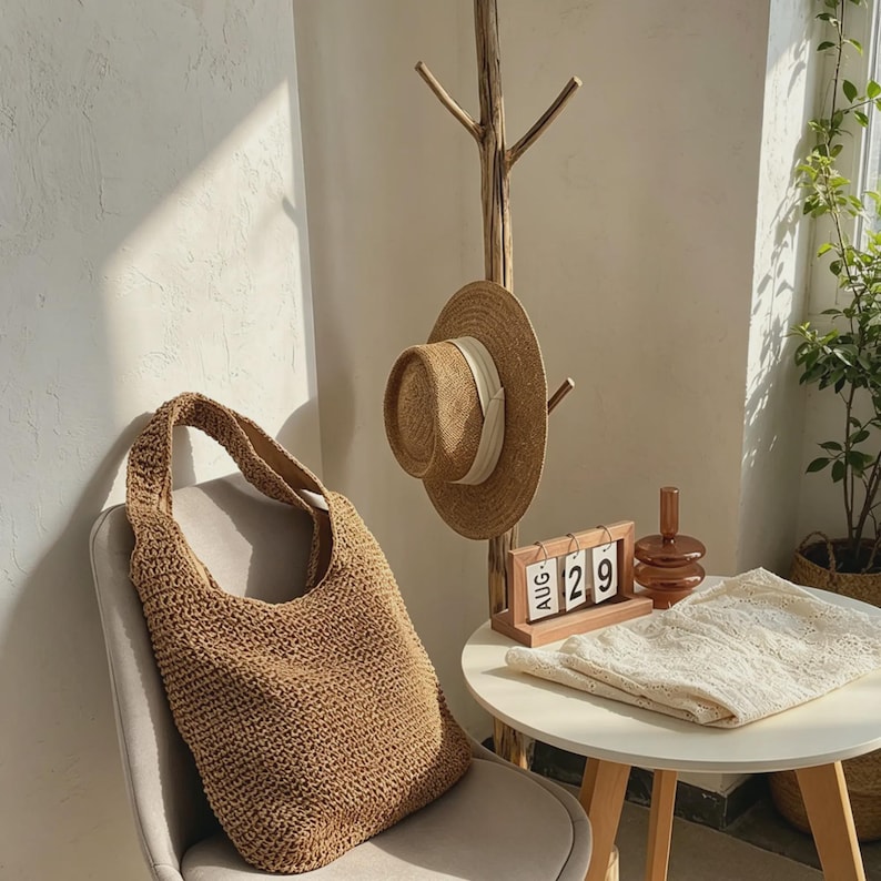 Puede incluir: Un bolso tote de paja tejida sobre una silla. Un sombrero de paja con una cinta blanca cuelga de un perchero de madera. Un peque&ntilde;o calendario de madera muestra el 29 de agosto. Un pa&ntilde;o de encaje blanco est&aacute; sobre una mesa.