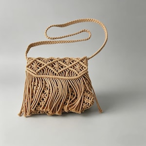 Puede incluir: Bolso bandolera de macramé beige con correa trenzada y flecos. El bolso presenta un patrón geométrico en la solapa y un cuerpo tejido. La correa es lo suficientemente larga para llevarla al hombro.