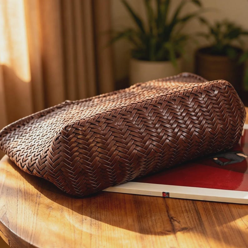 Peut inclure: Un sac &agrave; main marron tiss&eacute; de forme rectangulaire. Le sac est fait d'un mat&eacute;riau tiss&eacute; serr&eacute;, cr&eacute;ant une surface textur&eacute;e. Le sac repose sur une table en bois.