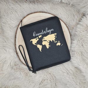 Könnte beinhalten: Schwarzer Reise-Dokumentenhalter mit goldenem Weltkarten-Design und dem Wort "Reiseunterlagen" in Schrift. Der Halter hat eine Handschlaufe und einen Reißverschluss. Der Halter liegt auf einer Holzscheibe.