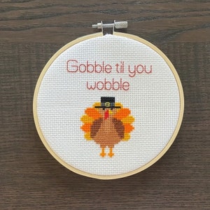 May include: A white embroidery hoop with a cross-stitch design of a turkey wearing a pilgrim hat. The text "Gobble til you wobble" is stitched above the turkey.
