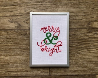 Puede incluir: Un bordado de punto de cruz enmarcado con tela blanca y marco plateado. El bordado presenta las palabras "merry & bright" en hilo rojo y verde.
