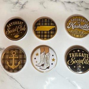 May include: Six circular buttons featuring various designs: one says Saturday Social Club, one shows a Tennessee map on plaid, one says Saturdays in Nashville, one features a gold anchor on plaid, one shows white cowboy boots, and one says Tailgate Socialite.