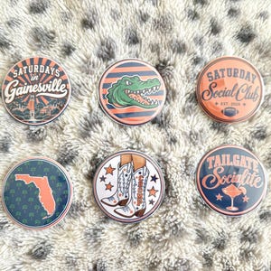 May include: Six circular pinback buttons featuring various designs in orange, blue, and white. Designs include text reading Saturdays in Gainesville, Saturday Social Club, Tailgate Socialite, a green alligator, a Florida map, and cowboy boots.
