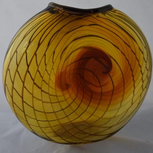 May include: A round, yellow glass vase with a dark brown, web-like pattern. The vase has a dark brown rim and a swirling, amber-colored center. The glass appears to be hand-blown, with a unique design.