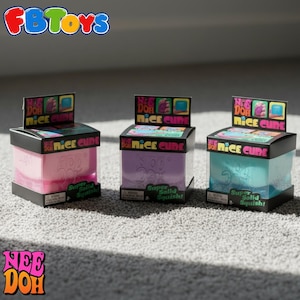 Puede incluir: Tres juguetes Nee Doh Nice Cube en rosa, morado y azul, cada uno en una caja negra con una pantalla superior colorida. Las cajas tienen el texto "Nice Cube" y "Super Solid Squish!". Los logotipos de FB Toys y Nee Doh son visibles.