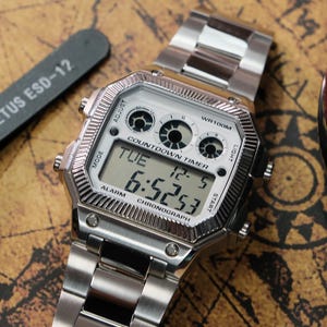 Puede incluir: Un reloj de pulsera digital plateado con una esfera blanca que muestra la hora y la fecha. El reloj tiene una correa de acero inoxidable y está sobre una superficie con forma de mapa. El texto "VETUS ESD-12" es visible.