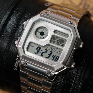 May include: A silver digital watch with a stainless steel band. The watch face displays the time as 9:23:48 PM, along with other digital readouts. The watch has a geometric design and is set against a dark background.