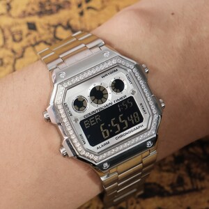 Op de afbeelding: Zilverkleurig digitaal horloge met een rechthoekige wijzerplaat en een zilver-gouden band. Het horloge toont de tijd 6:55:48 en heeft een countdown timer en chronograaf functies. Het horloge is versierd met kleine, heldere steentjes.