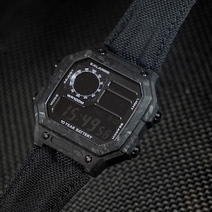 Op de afbeelding: Een digitaal horloge met een vierkante behuizing met zwart marmerpatroon en een zwarte stoffen band. De wijzerplaat toont de tijd 15:49:58, de tekst 5 ALARMS, WR100M en 10 YEAR BATTERY, samen met labels voor de zijknoppen: ADJUST, MODE, LIGHT en SEARCH.