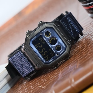 Op de afbeelding: Een digitaal polshorloge met een donkergrijze metalen kast en een blauwe stoffen band. De wijzerplaat toont de tijd, 12:55:35, en heeft verschillende functie-indicatoren. Het horloge is waterdicht tot 100 meter.