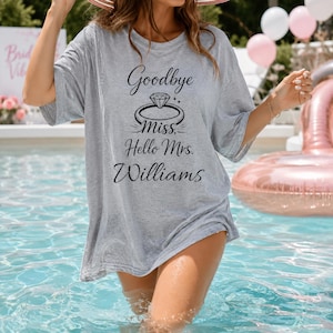 Puede incluir: Una camiseta gris jaspeado de manga corta con un gráfico negro de un anillo de diamantes y el texto "Goodbye Miss, Hello Mrs. Williams" impreso en una combinación de fuentes script y serif centrado en la parte delantera.