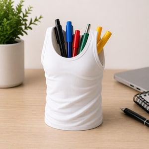 Peut inclure: Un organiseur de bureau blanc en forme de débardeur rempli de stylos. L'organisateur est fait d'un matériau blanc uni et a une texture côtelée. Les stylos sont noirs, bleus, rouges, verts et jaunes. L'organisateur est sur un bureau en bois.