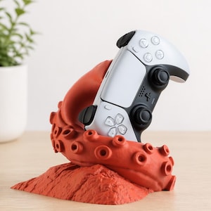 Könnte beinhalten: Ein weiß-schwarzer Videospiel-Controller ruht in einem roten, tintenfischförmigen Ständer. Der Ständer hat eine strukturierte Oberfläche und Saugnapfdetails. Der Controller hat Knöpfe und Richtungspads.