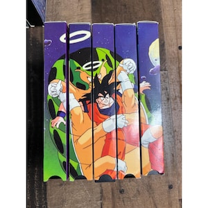 May include: A stack of five VHS tapes featuring colorful illustrations of characters from the Dragon Ball Z anime series. The spines are purple with character art and the series logo. The artwork depicts action scenes with characters in orange and blue.