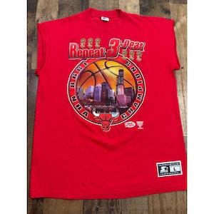 May include: Red sleeveless t-shirt featuring a graphic commemorating the 1998 NBA Champions. The design includes a basketball, cityscape, and the text "Repeat 3-Peat". The Chicago Bulls logo is also present.