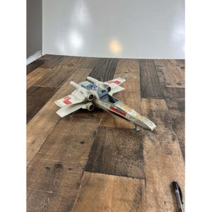 May include: A toy X-wing fighter in off-white with red and orange accents. The model has four engines, wings, and a cockpit. The toy is made of plastic and sits on a wooden surface. A pen is in the lower right corner.