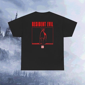 May include: A black short-sleeve t-shirt featuring the red text RESIDENT EVIL above a red framed graphic of a sitting dog. Below the graphic is a small red and white umbrella symbol. The design is centered on the back of the garment.