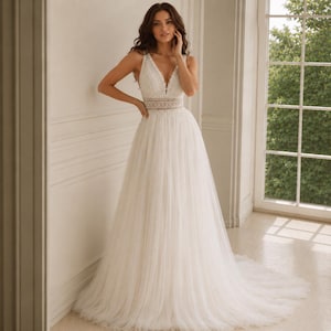 May include: A floor-length white gown featuring a sleeveless bodice with a deep V-neckline, intricate lace detailing, and a defined waistline. The skirt is constructed from layers of sheer tulle, creating a full, flowing silhouette that extends into a train.