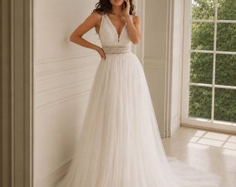 May include: A floor-length white gown featuring a sleeveless bodice with a deep V-neckline, intricate lace detailing, and a defined waistline. The skirt is constructed from layers of sheer tulle, creating a full, flowing silhouette that extends into a train.