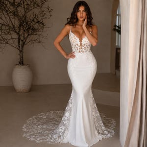May include: A floor-length white mermaid dress featuring a deep V-neckline, thin spaghetti straps, and a bodice adorned with intricate floral lace embroidery. The skirt is made of smooth fabric with lace detailing along the hem and a long train.