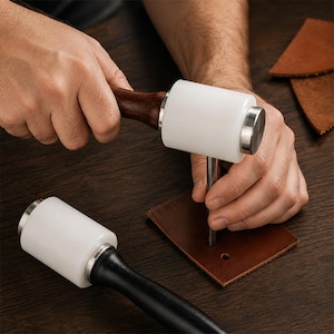 May include: Two leather crafting mallets featuring cylindrical white heads and dark wooden handles. One mallet is being used to strike a metal tool positioned over a small square piece of brown leather, while the second mallet rests nearby.