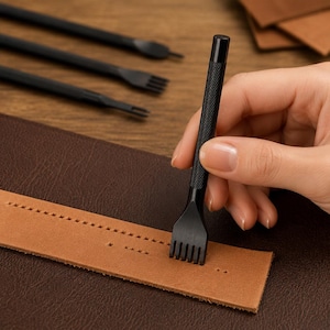 Peut inclure: Un outil de piquage en cuir en métal noir avec une poignée texturée et moletée. L'outil est positionné pour percer une rangée de trous dans une bande de cuir beige. D'autres outils de maroquinerie noirs sont visibles en arrière-plan.