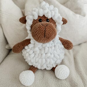 May include: A plush stuffed animal shaped like a sheep, featuring a brown face and limbs with a textured white wool body. The sheep has two black plastic eyes, a stitched nose and mouth, and a fluffy white head covering that frames the face.
