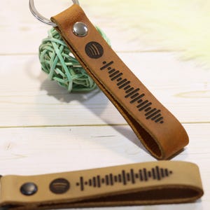 May include: Two leather keychains with a Spotify code design. The keychains are brown and tan with a metal key ring. The Spotify logo is visible on the keychain. The keychains are on a white wooden surface.