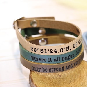 May include: A stack of three leather bracelets in tan, green, and brown. Each bracelet is engraved with text and coordinates. The bracelets are stacked on a wooden slice, with a phone and decorative ball in the background.