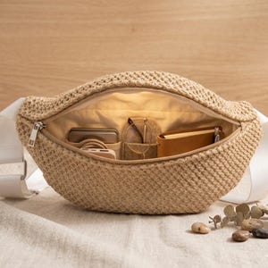 May include: A beige woven bum bag with a white strap, open to show a light-coloured interior. Inside are a phone, glasses, a small notebook, and other items. The bag is on a neutral surface.