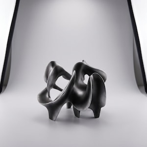 May include: A dark gray abstract sculpture with a flowing, organic form. The sculpture has a matte finish and appears to be made of a solid material. The design features curves and negative space, creating an intriguing visual effect. The artwork is displayed on a light gray surface.