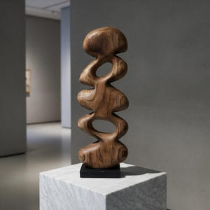Può includere: Una scultura astratta in legno con una serie di forme arrotondate e interconnesse e spazi aperti. La scultura è esposta su un piedistallo di marmo bianco con base nera, su uno sfondo neutro da galleria. La venatura del legno è visibile.