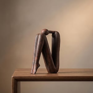 Pode incluir: Uma escultura abstrata castanha de uma pessoa sentada, com as pernas levantadas e a cabeça apoiada nos joelhos. A escultura tem uma superfície texturizada e é exibida numa mesa de madeira.