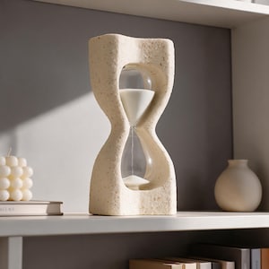 May include: An hourglass featuring a textured, speckled beige stone-like frame with a curved, organic shape. The central glass bulb contains fine white sand, with the top bulb partially filled and the bottom bulb holding a small amount of sand.