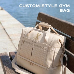 Könnte beinhalten: Eine beige Sporttasche mit gesteppter Vordertasche, goldfarbenen Reißverschlüssen und einem Tragegriff. Die Tasche trägt den Text "CUSTOM STYLE GYM BAG" oben und "MAELSTROM" auf der Vorderseite sowie ein goldenes Sticklogo mit den Buchstaben A und L.