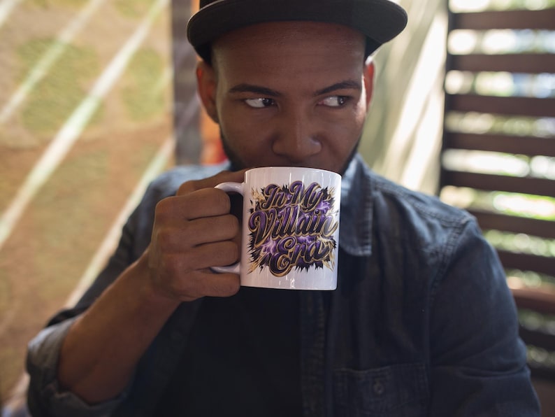 May include: White ceramic mug featuring the text "In My Villain Era" in purple and gold lettering. The mug has a black handle and a black and purple design. The mug is held by a person.