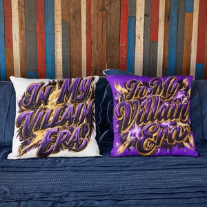 May include: Two decorative cushions with the text "In My Villain Era" in purple and gold lettering. One cushion is white, the other is purple. The cushions are on a blue bed with blue cushions and a colourful wooden background.