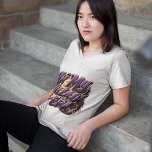 May include: A light grey V-neck t-shirt featuring the text "IN MY VILLAIN ERA" in purple and gold. The person is sitting on concrete steps. The shirt is a casual wear item.