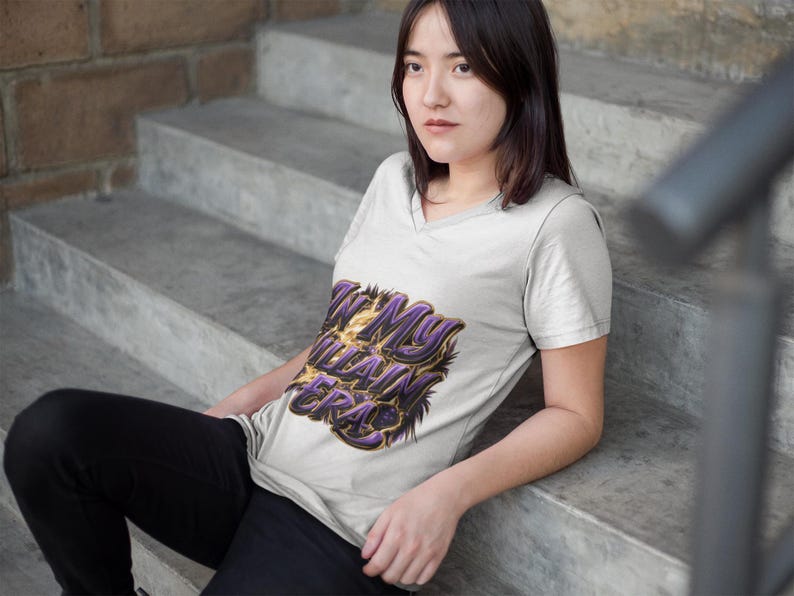 May include: A light grey V-neck t-shirt featuring the text "IN MY VILLAIN ERA" in purple and gold. The person is sitting on concrete steps. The shirt is a casual wear item.