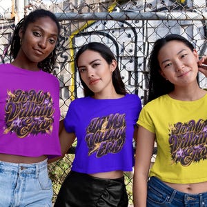 May include: Three women wearing crop tops in purple, blue, and yellow. Each top features the text "In My Villain Era" in a stylized design with gold and purple accents. They are standing in front of a chain-link fence.