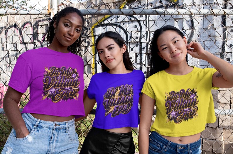May include: Three women wearing crop tops in purple, blue, and yellow. Each top features the text "In My Villain Era" in a stylized design with gold and purple accents. They are standing in front of a chain-link fence.