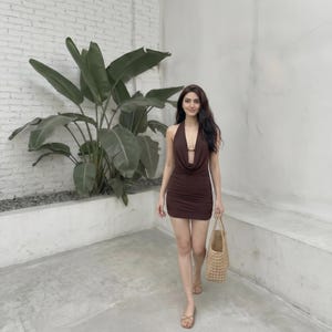 May include: A brown halter dress with a low neckline. The dress is ruched and form-fitting. A woven straw tote bag and sandals complete the look. A large green plant is in the background.